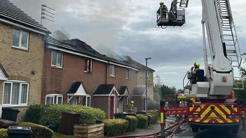 Fire crews and fire engine attending to a fire on a residential street, smoke can be seen coming from the roof of one of the homes