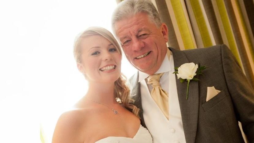 A young woman on her wedding day wearing a white dress smiling with her older father, who is dressed in a suit