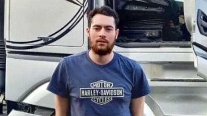 Gregory Peter Newcombe wearing a blue t-shirt that said: "Motor Harley-Davidson Cycles" and has black hair and a black beard. He is also stood in front of a lorry with one of the doors open