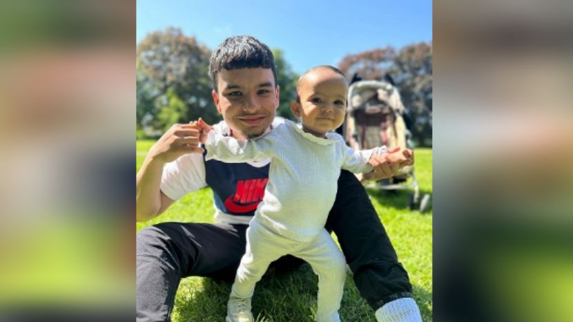 Mr Qureshi, wearing a white and blue Nike T-shirt with a Nike logo on it, sitting on grass with his baby daughter as she stands up and they smile at the camera