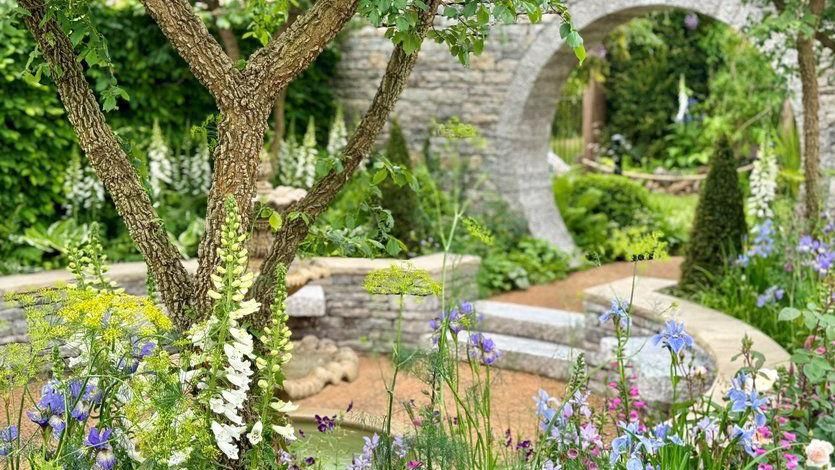 Entry to the garden is under a circular arch, heading down two steps to the middle where the water feature sits. It is surrounded on all sides by flowers and trees. 