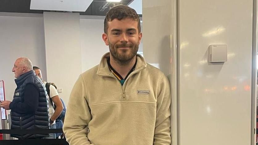 Luke Watters is standing in an airport, leaning against a pillar, wearing a cream fleece. Some people can be seeing queuing in the background 