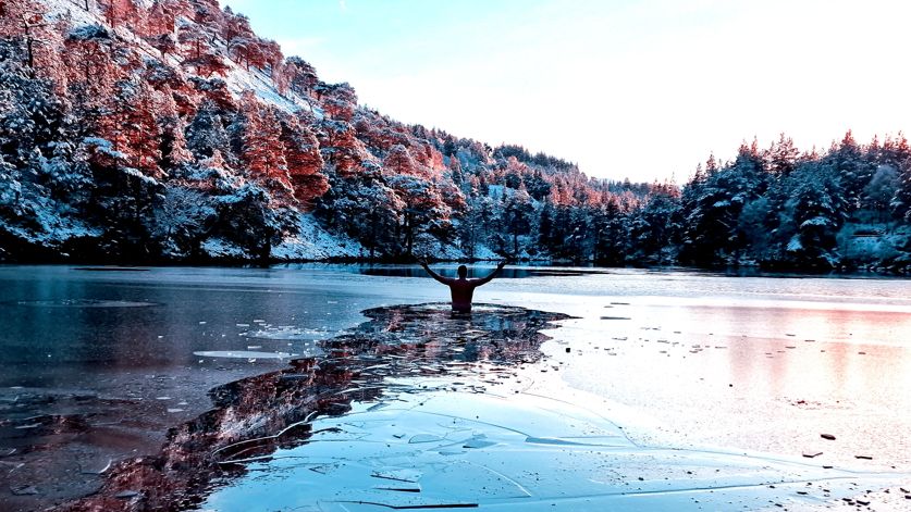 Adam Baker standing in icy waters with his hands raised in the air. There is a frozen layer on the surface of the water, which he has broken through and left a trail. Snow-covered trees are visible on the banks of the lake