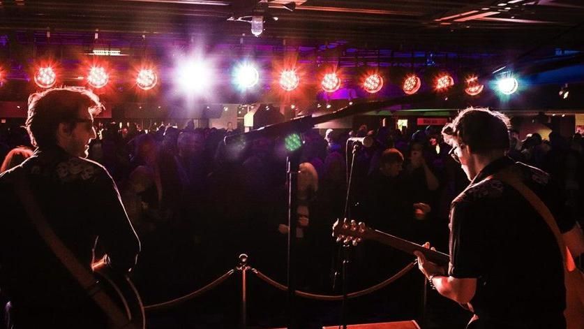 Two men playing guitars on a stage with coloured spotlights on them performing in front of a crowd