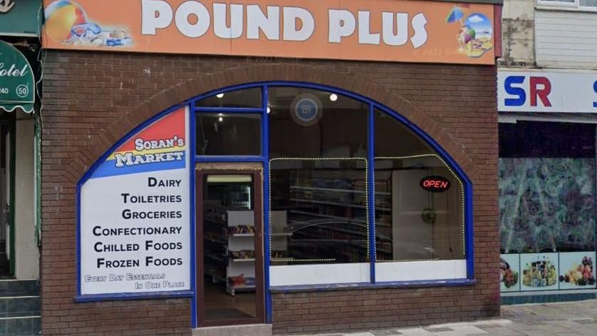 A food shop on Coronation Street in Blackpool with an orange sign above it saying Pound Plus and a blue lined window saying Soran's Market - selling Dairy, Toiletries, Groceries - a neon red sign says Open in the corner