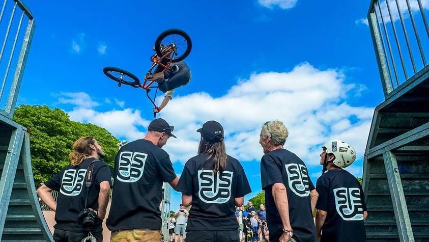 Members of the Swanage Skatepark Community Project wearing branded t-shirts and watching as someone on a BMX flies over head after rising up a ramp