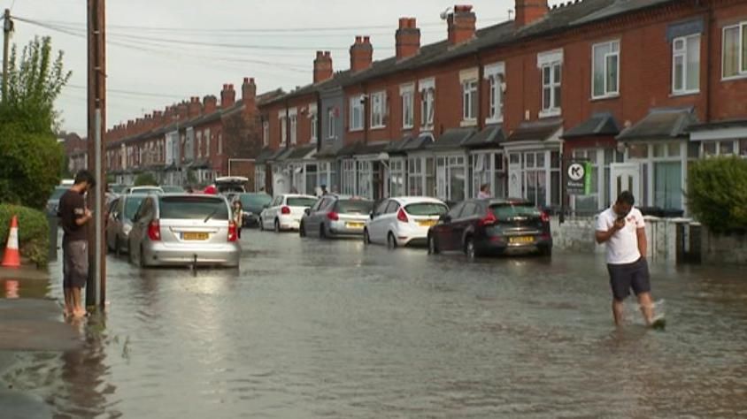 A flooded residential road in Sparkhill, Birmingham, showing a row of terraced redbrick houses and parked cars under inches of water. A man in shorts standing ankle deep in water is on the left with another in a white t-shirt walking down the middle of the road.