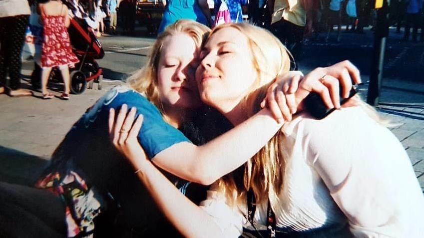 Gaia and Ms Pope-Weidemann hugging and smiling. they are sitting down in a street. Gaia is wearing a blue top and Ms Pope-Weidemann is wearing a white jumper. Both have long blonde hair