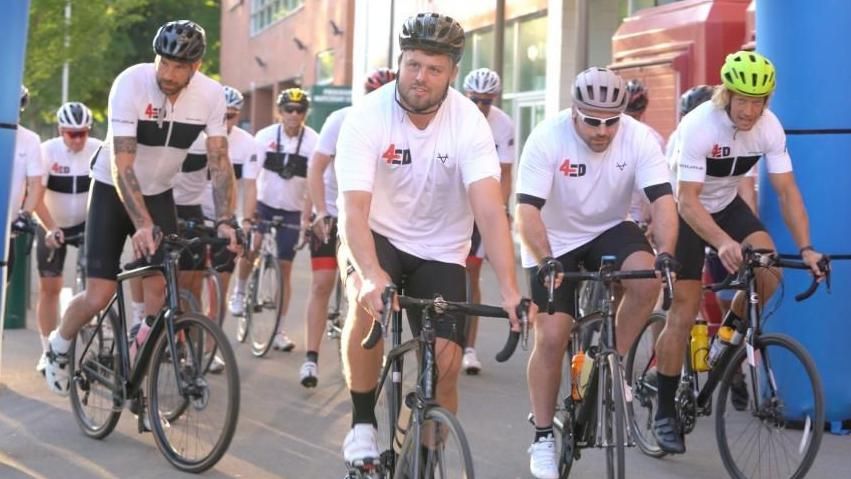 Mr Slater at the forefront of the picture riding a bike through the inflatable finish line of the three day cycle. He is joined by around nine others, who are also cycling through the finish line behind him. They are all wearing white t-shirts that say '4Ed' and black cycling shorts