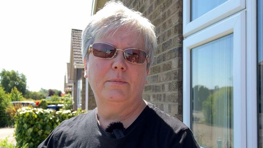 Paula De Vries, with short, grey hair, is wearing sunglasses and a black t-shirt as she stands in her front garden