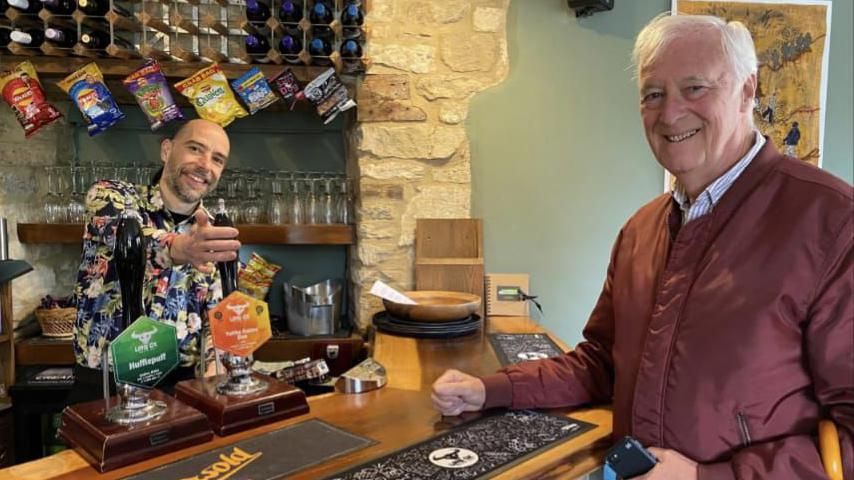 Craig Tipper pouring a pint for a man who is wearing a red coat
