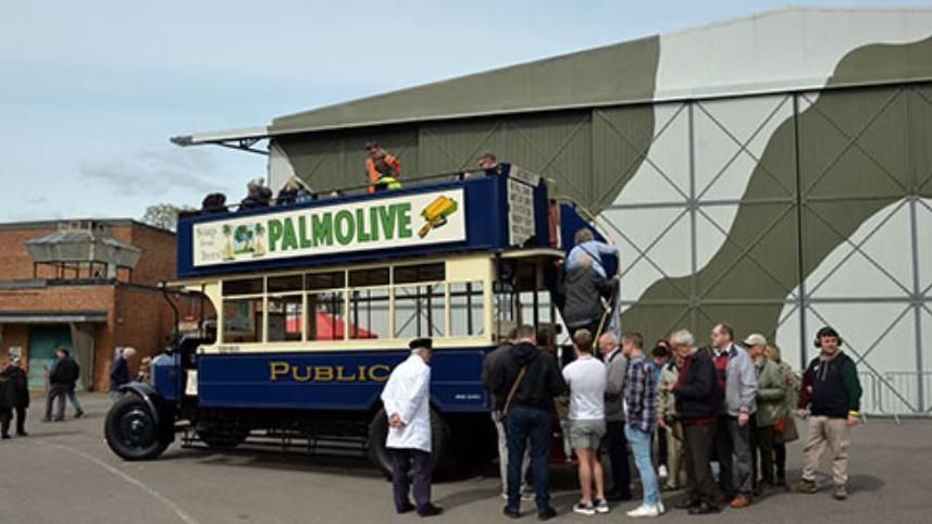 A heritage open top bus painted in royal blue with vintage advertising hoardings is parked outside a green hangar with spectators queuing to board it