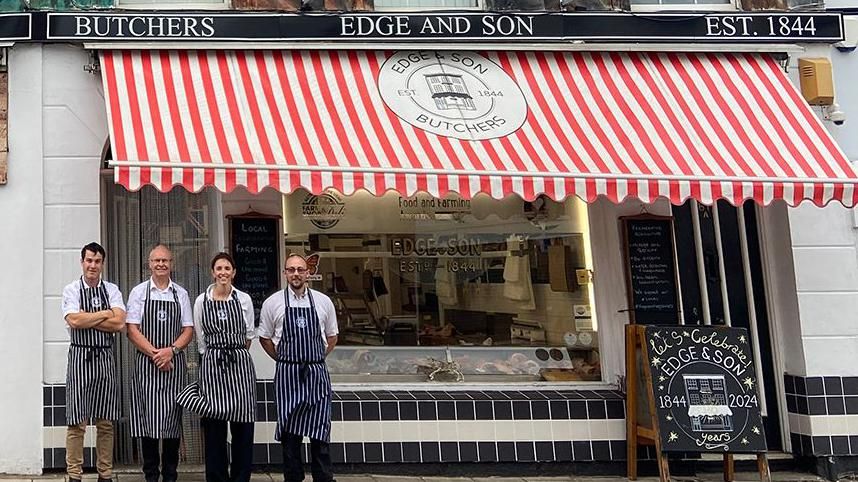 Butchers outside the Edge & Son shop in New Ferry. The shop's exterior is decorated with blue and while square tiles, while a red and white canopy with the Edge & Son logo hangs over the window. There's a blackboard outside the door that reads 'Let's celebrate'.