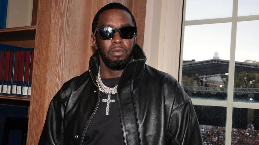 Sean "Diddy" Combs pictured wearing black sunglasses, a black leather jacket, a jewelled chain with a cross on it and a black T-shirt. 