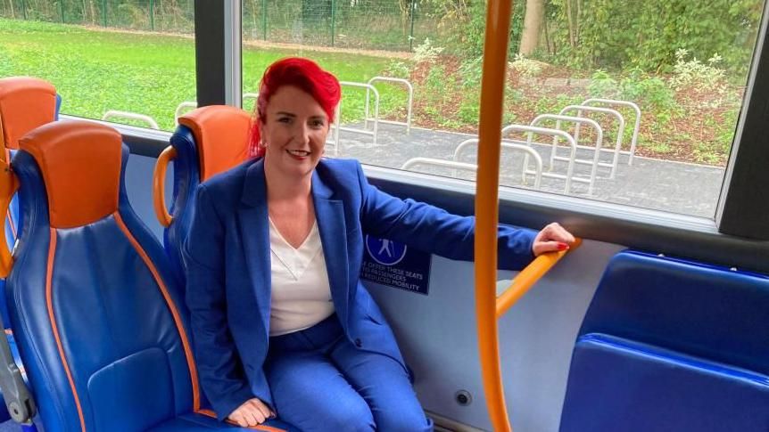 Louise Haigh sitting on bus, wearing a blue trouser suit and a white shirt. She is holding onto a yellow handrail