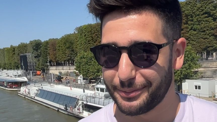 A young man with close-cropped dark hair and stubble wearing dark sunglasses and a pale pink t-shirt stands by the River Seine