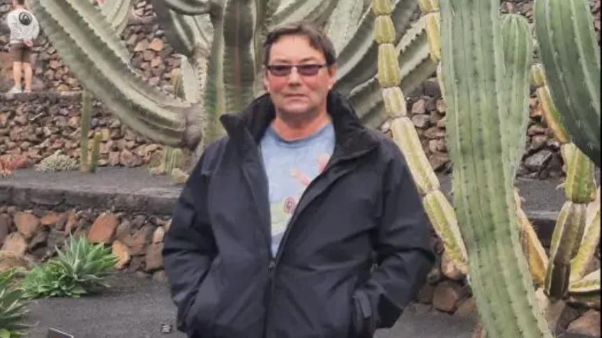 Franklin Ingram wearing sunglasses, a blue graphic t-shirt and a black zip up jacket. He is standing in front of large cacti and brown crumbly rubble