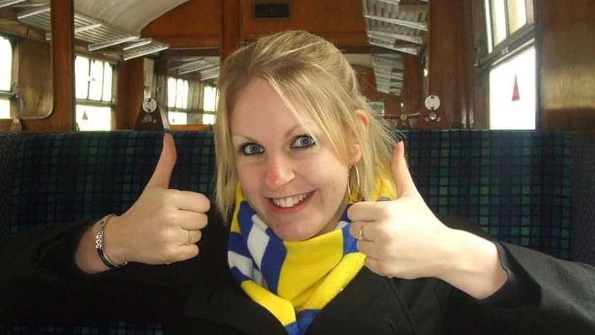 Gemma Sisley is sitting on a train, smiling at the camera with both thumbs up. She is wearing a black jacket and yellow and blue striped scarf. 