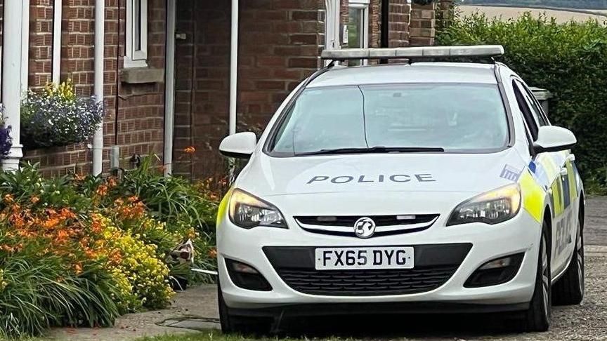 A Lincolnshire Police car is parked outside Christine Emmerson's home in West Ashby on a gravel driveway. Plants and flowers are growing around the front of the house and there is a white front door with white window frames.