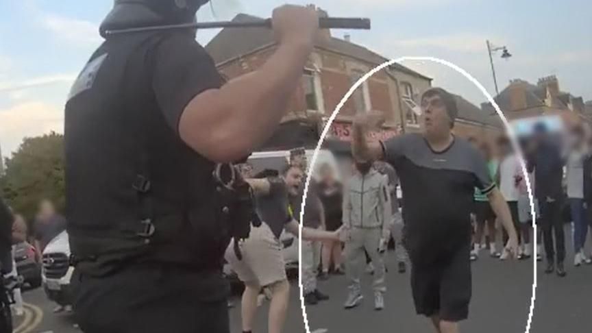 A still from police footage showing two men gesticulating at a police officer with a crowd behind them