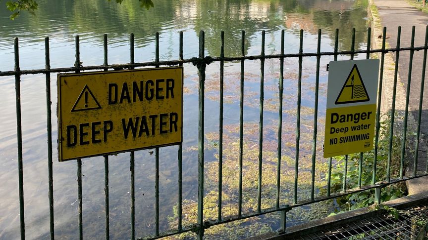 Yellow and black signs, reading "danger deep water", are fixed to a metal fence, in front of a wide body of water.