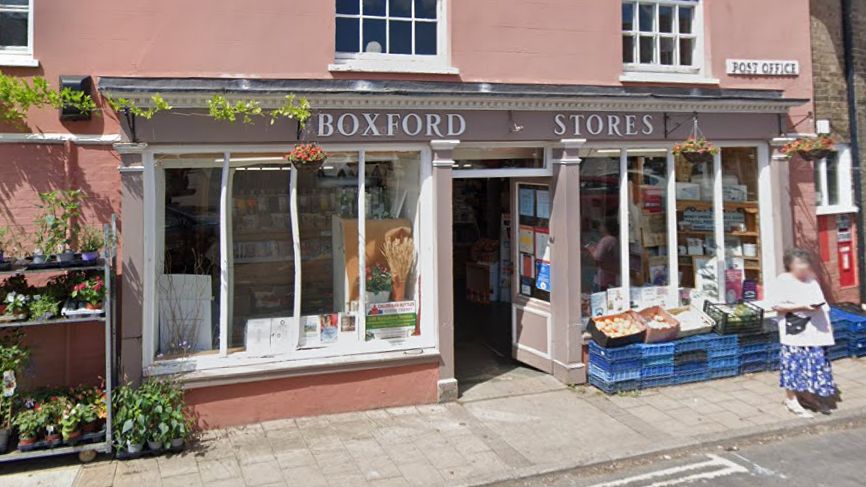 Outside Boxford Stores Post Office. The shop exterior is dusky pink with large windows and fruit, vegetables and plants in boxes outside. There is a red post box built into the wall next to the shop. 