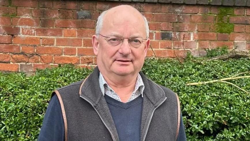 Andrew Reid standing in front of a bush, wearing glasses, a gilet/bodywarmer over a blue jumper and checked shirt.