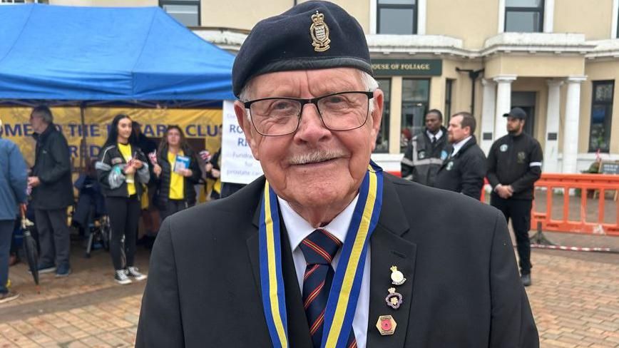 Smiling veteran Eric Howden in front of a crowd wearing a hat with military badge and medals pinned to his lapel