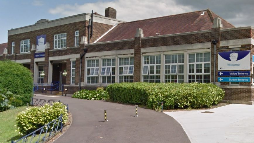 The entrance driveway to Hathaway Academy. There is a lawn to the left and bushes in front of the school building, which has many windows and is brick.