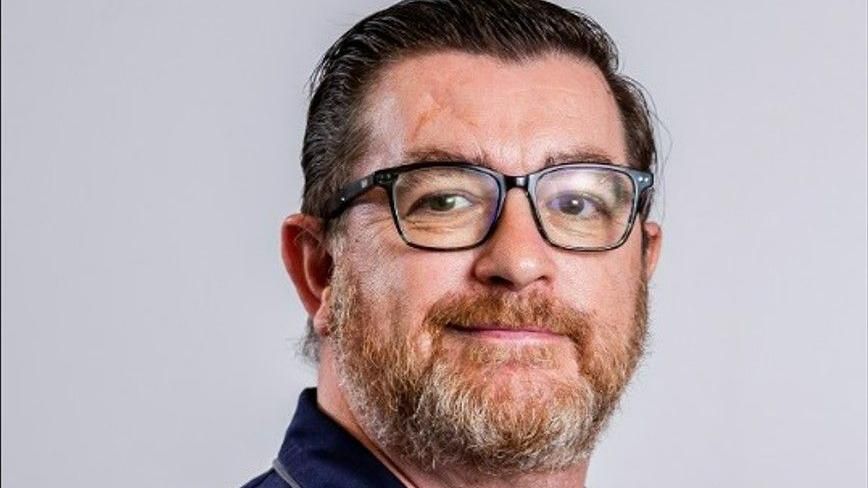 Paul Brittain, a man with a beard and brown hair and glasses, wearing a polo neck T-shirt