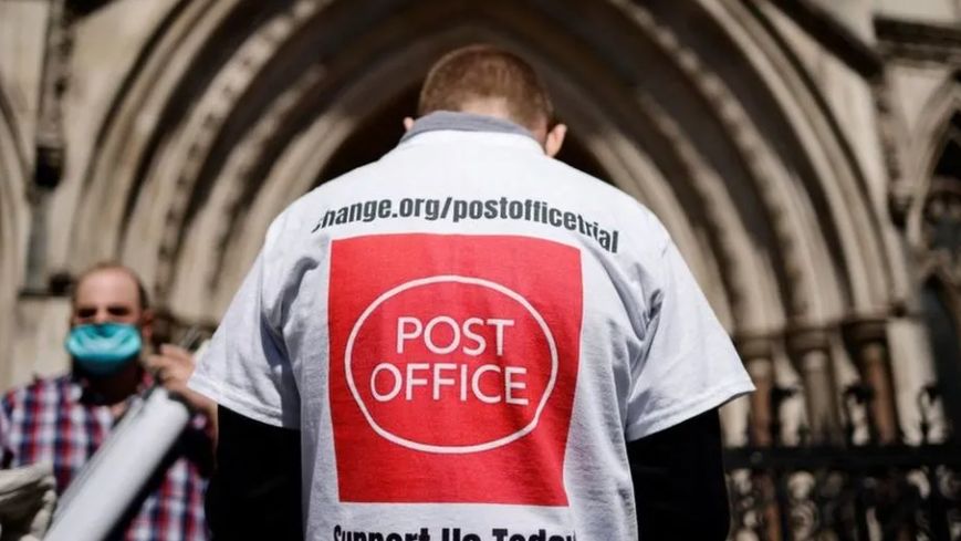 A campaigner outside court from behind, wearing a t-shirt emblazoned with a Post Office logo and slogan