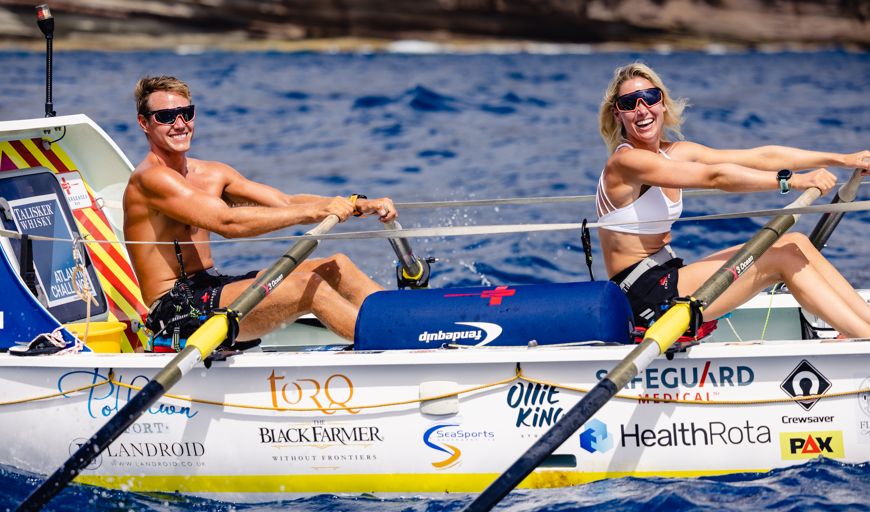 Adam Baker and Charlie Fleury rowing across the Atlantic. They are sitting in a customised boat, in the sunshine, both smiling at the camera