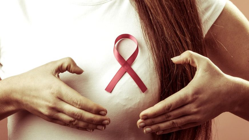 Mujer joven seálando a uno de sus senos donde lleva el lazo representativo de la lucha contra el cáncer de mama.