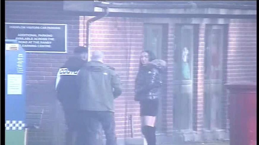 Stephanie Heaps stands outside prison wearing over the knee black heeled boots, a black skirt and a coat with furry hood, with two men seen to the left of her, their faces not visible