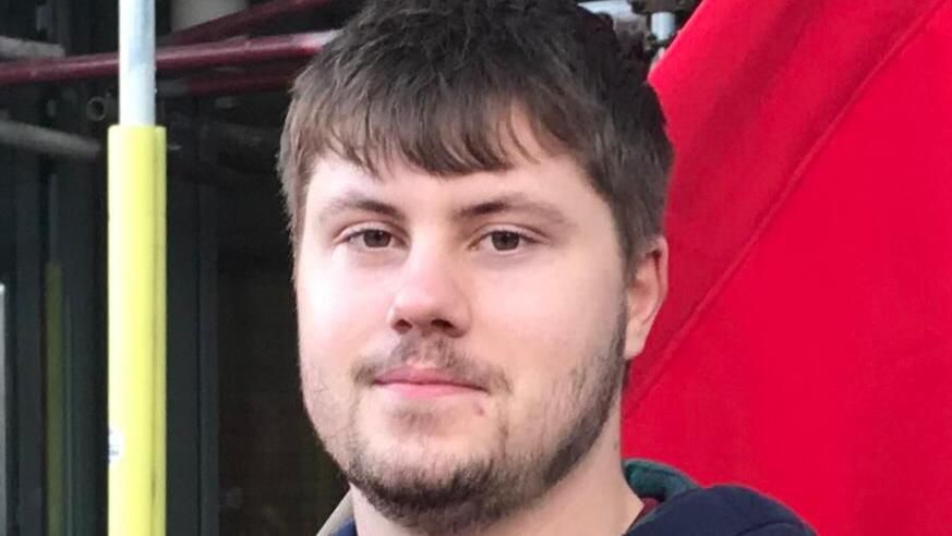 Fraser McGuire. Fraser is a 20-year-old man with short brown hair, a short brown beard and brown eyes. He's pictured outside in front of a red background 