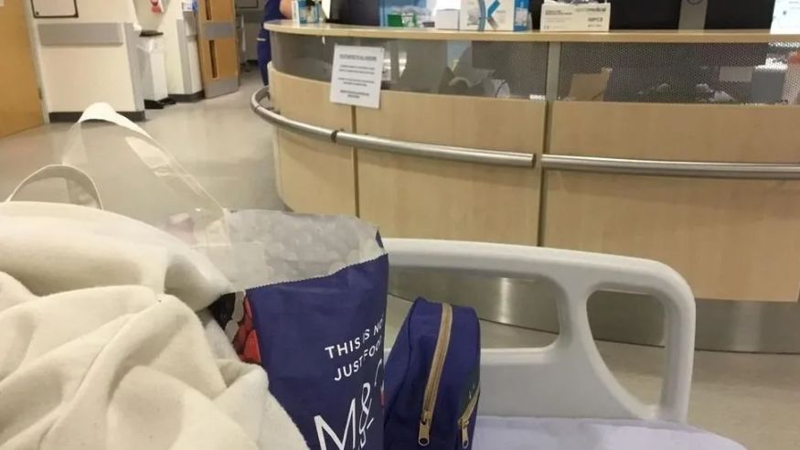 A view from the bed of a patient being treated in a corridor at the hospital. In the foreground is a shopping bag and a blanket. In the background is what appears to be a ward reception desk