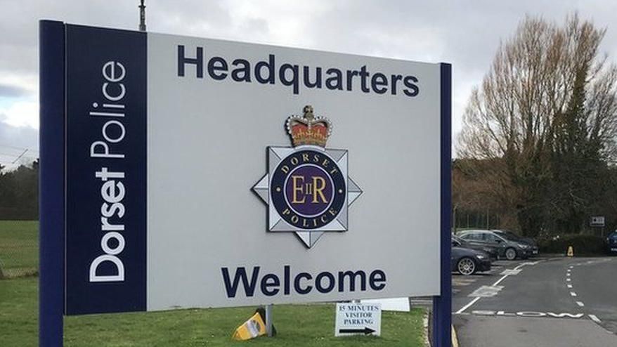 Sign outside Dorset Police HQ bearing the force's badge. The blue and white sign reads: Dorset Police Headquarters Welcome