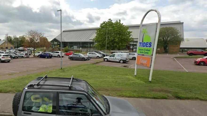 View from the road of Tides leisure centre, with cars parked in front, and a green sign next to the road