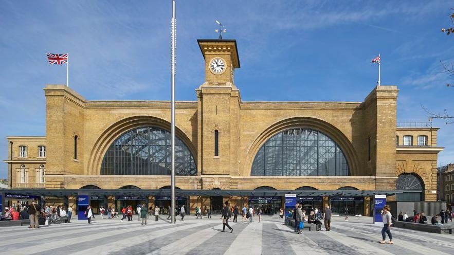 Outside King's Cross railway station in London, showing a yellow brick-built building, with two very large glazed arches into the station, a tower in the middle with a clock, and people walking in front of the station on a grey striped public square