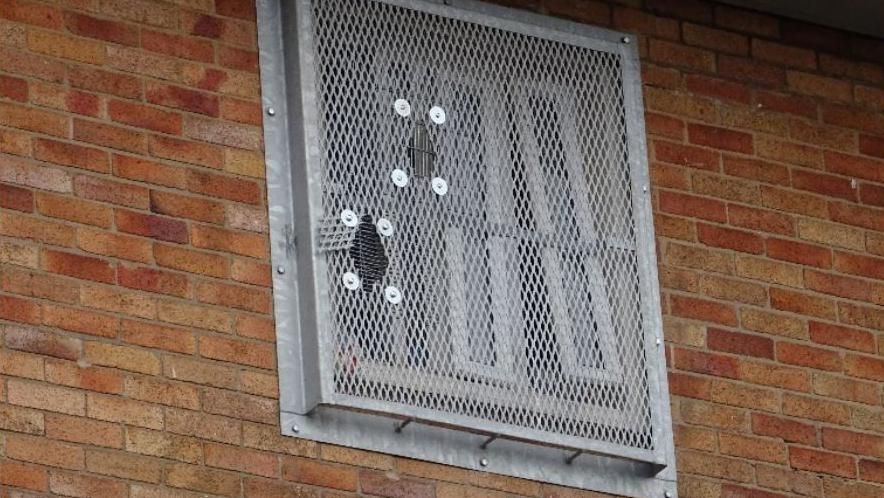 A damaged prison window with holes in the mesh guard covering the site.