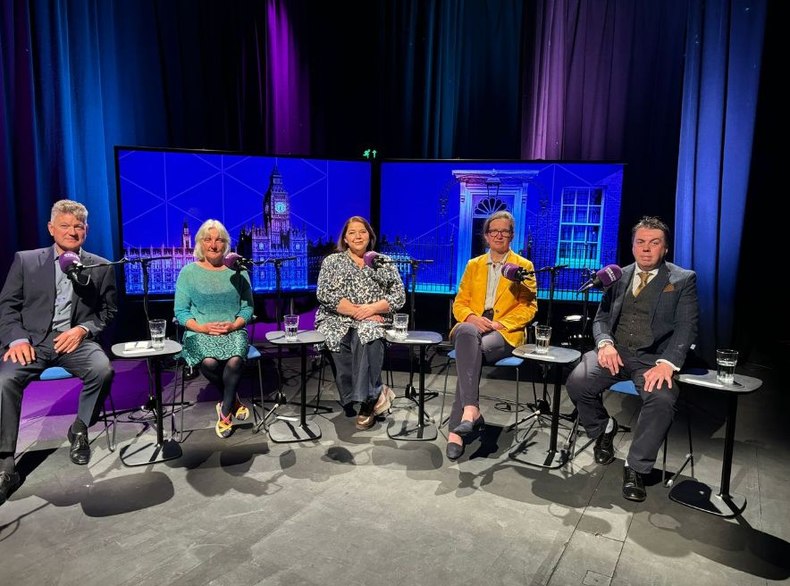 The candidates sitting in a row. They each have a table with a glass of water on to their left and a microphone. There are curtains and branded screens behind them.