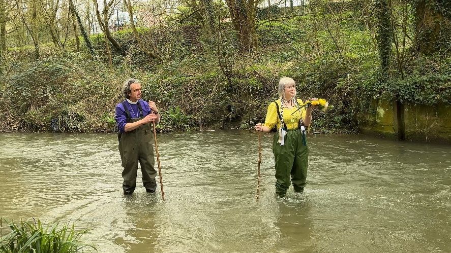 The Bishop of Ramsbury, Andrew Rumsey, and Tara a flower maiden from Stroud giving the blessing whilst standing in the river Marden wearing waders