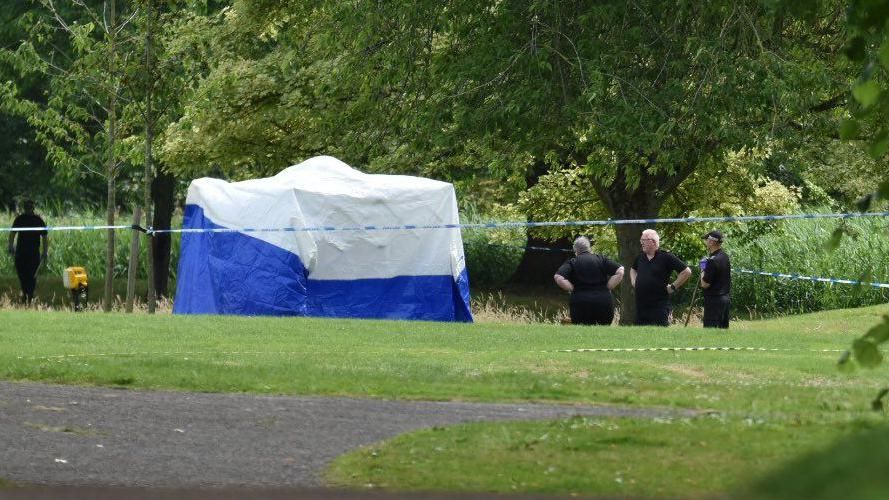 Three people in black uniforms standing by a white and blue forensics tent on the grass, which is surrounded by police tape