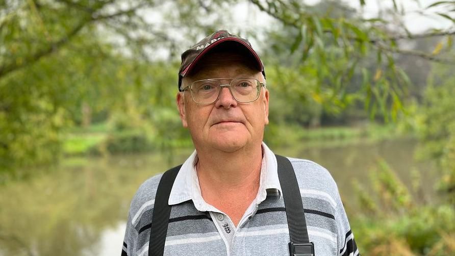 Stephen Bell wearing glasses and a cap stands in front of the lake looking into the camera