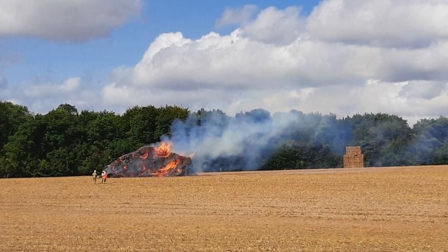 A burning haystack in a field with smoke rising from it. There is a second undamaged haystack in the background