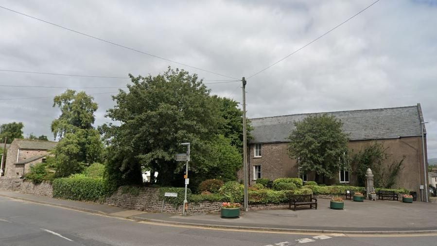 View of a small public space on a street corner, with a low wall and a hedge behind two seating benches.