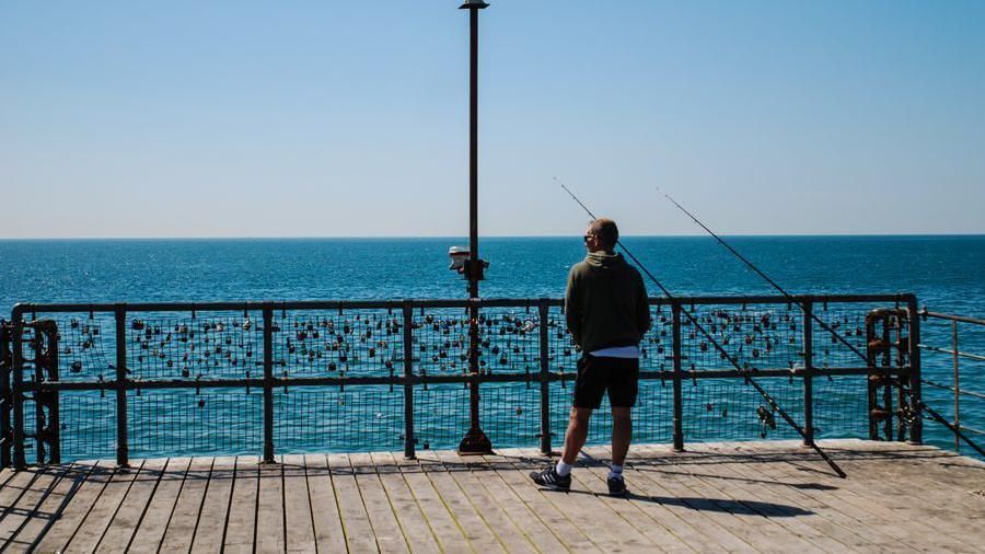 A man standing on a pier in front of the sea with two fishing poles beside him 