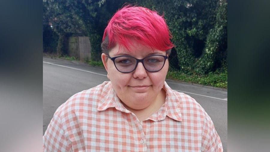 A woman smiling, she has pink, short hair and is wearing a checked pink shirt.