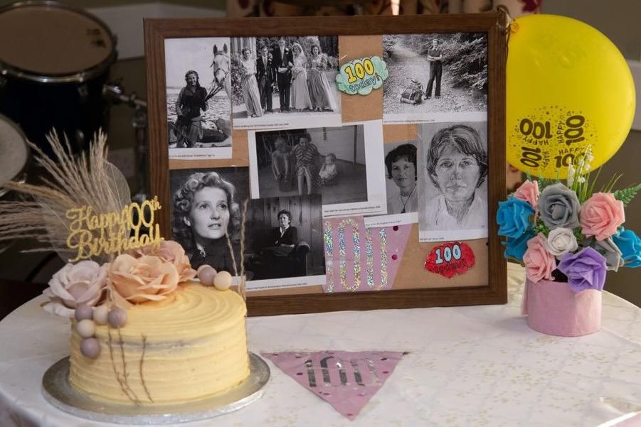 A montage of black and white photos of Jean, next to a 100th birthday cake with flowers on it and a "Happy 100th Birthday" topper, as well as a yellow balloon and flowers