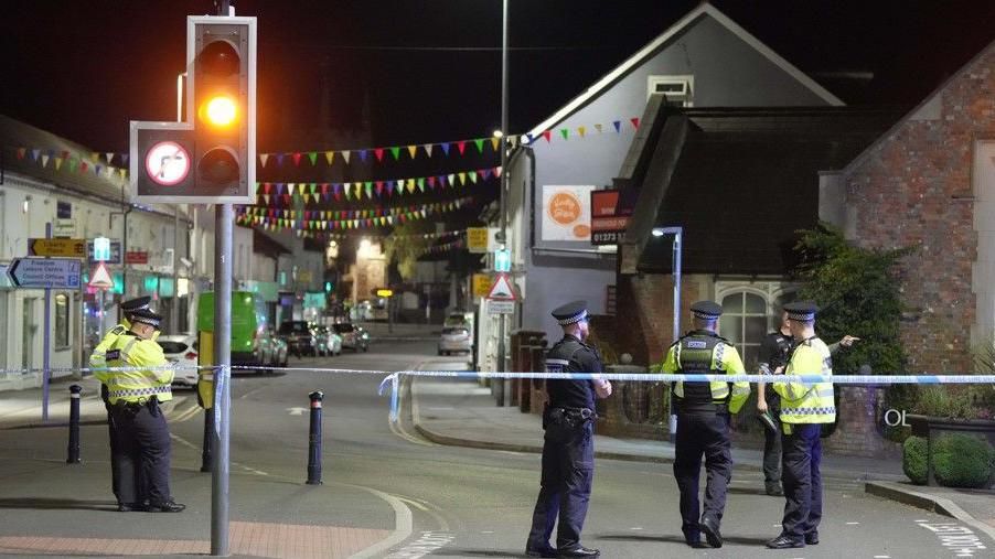 The town centre at night with police tape and several police officers, some wearing high-vis jackets.